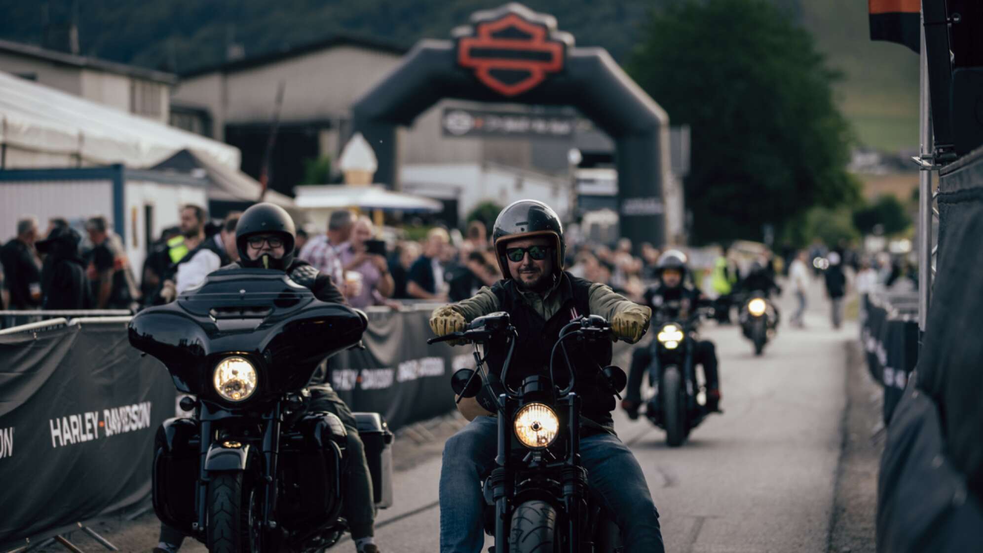 Motorradfahrer auf Harley-Davidson-Maschinen fahren bei den Bike Days Willingen 2026 durch eine abgesperrte Straße. Im Vordergrund sind zwei Biker mit Helmen und Schutzkleidung zu sehen, im Hintergrund jubeln zahlreiche Zuschauer am Straßenrand. Über dem Eingang ist ein großes Harley-Davidson-Logo zu erkennen, dahinter Berge und Veranstaltungszelte. Harley Days Willingen - Motorrad-Event und Biker-Treffen im Sauerland