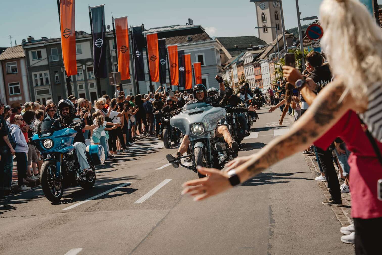 Harley-Davidson-Parade mit zahlreichen Motorrädern fährt durch eine von Publikum gesäumte Straße in Kärnten, Österreich, während der European Bike Week am Faaker See – Zuschauer jubeln und filmen den Korso.