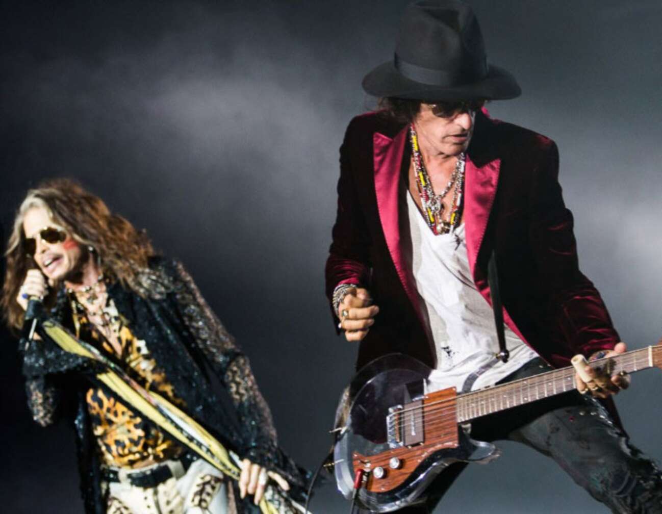 Joe Perry und Steven Tyler stehen in Bühnenpose, vor grauem Hintergrund