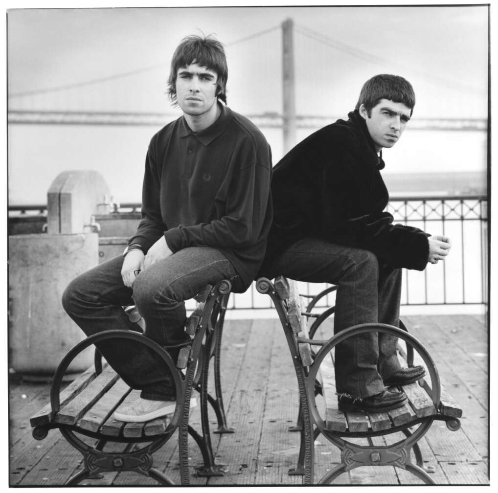 Noel und Liam Gallagher sitzend auf Bank, mit ernstem Blick