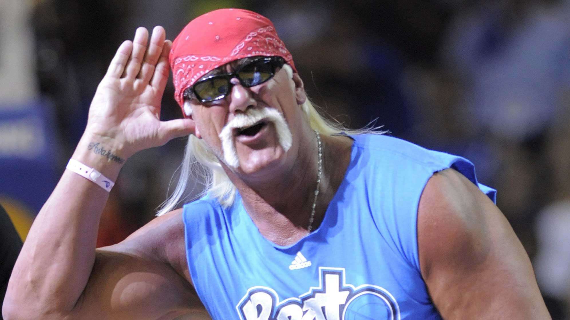 Hulk Hogen hebt die Hand an sein Ohr. Er trägt ein rotes Kopftuch, eine Sonnenbrille und einen Schnauzbart