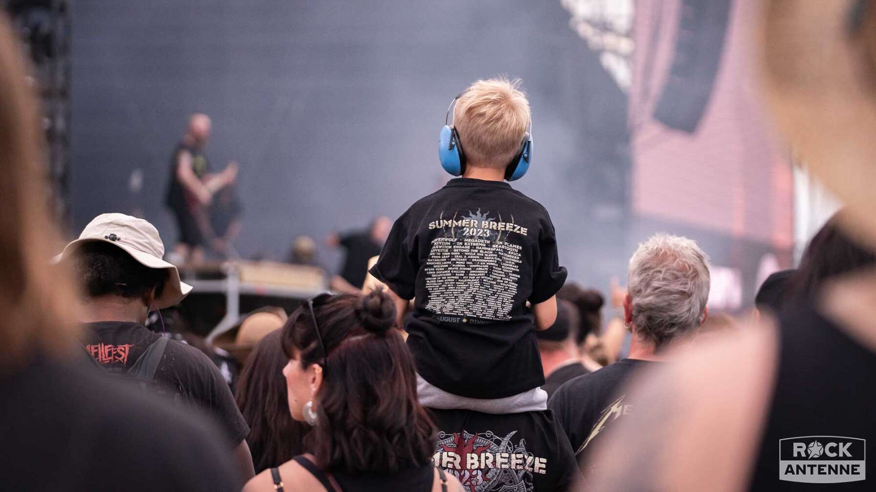 Ein kleiner Junge mit Kopfhörern sitzt auf den Schultern einer erwachsenen Person vor der Bühne des Summer Breeze Open Airs.