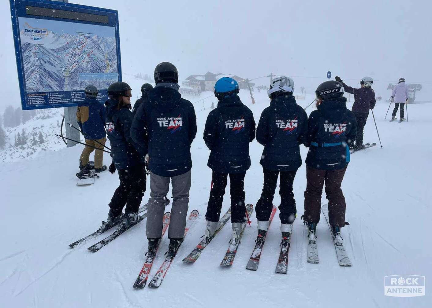 Foto der ROCK ANTENNE Tagesskifahrt Rock the Mountain 2023 in die Weltcuparena Zauchensee - Eine Gruppe Skifahrer von hinten fotografiert mit "ROCK ANTENNE Team"-Aufdruck auf der Jacke
