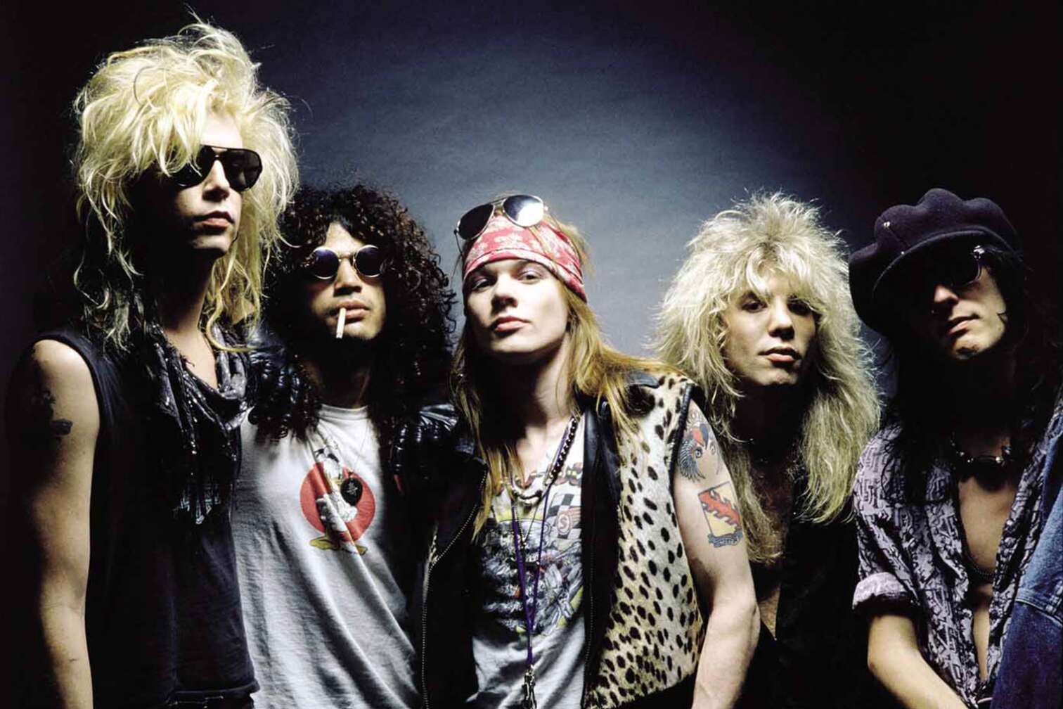 Guns N' Roses posieren mit Sonnenbrillen, Kippe und Bandana vor dunklem Hintergrund