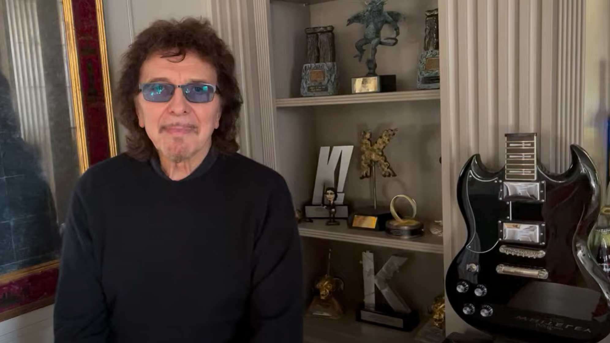 Screenshot des Neujahrs-Videos von Tony Iommi auf YouTube, Tony Iommi sitzt vor der Kamera, im Hintergrund ein Regal mit verschiedenen Statuen sowie eine Gitarre