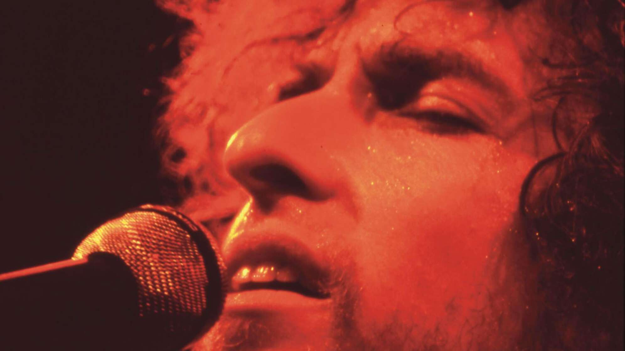 Bob Dylan singt mit geschlossenen Augen in rotem Scheinwerferlicht